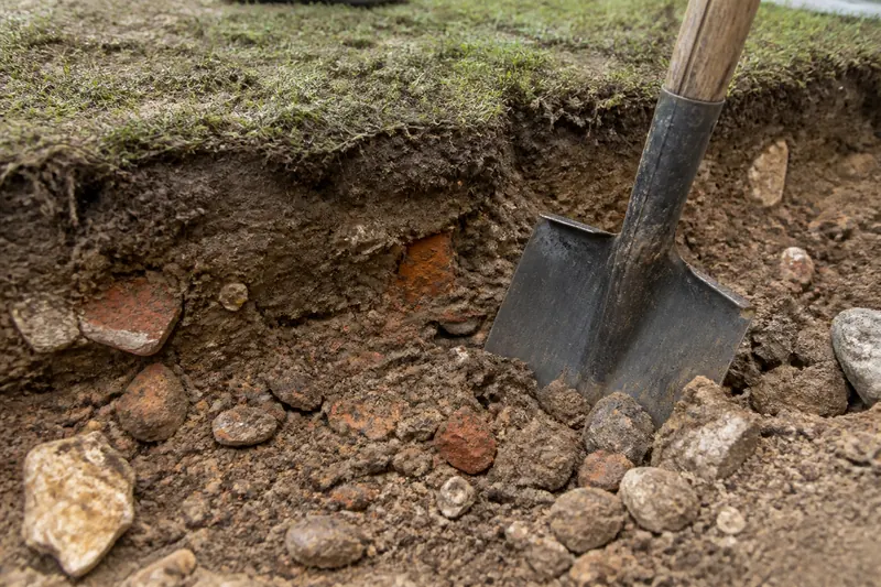 A spade cutting into clay and rubble under thin turf